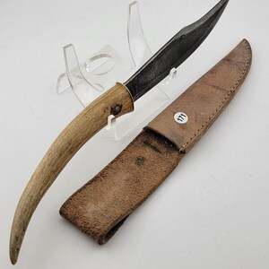 May include: A fixed-blade knife with a curved, textured tan handle and a dark, pointed steel blade. The knife is accompanied by a brown leather sheath featuring a small white circular sticker with the black number 171 printed on it.