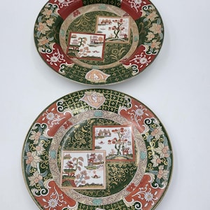 May include: Two round ceramic plates featuring a dark green and red border with floral patterns. The centre of each plate displays two square panels with landscape illustrations of trees and buildings against a gold and green background.
