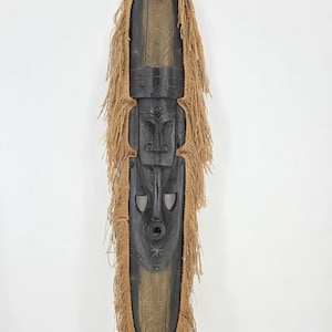 May include: A tall, elongated wooden mask featuring a dark black central face with carved facial features and light brown natural fibre fringe along the sides. The top and bottom sections of the mask are carved with light-coloured geometric patterns.