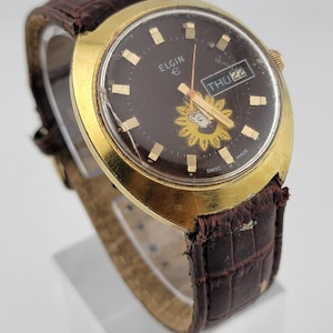 May include: Vintage Elgin watch with a brown face, gold-tone case, and a brown leather band. The watch face features gold markers, hands, and a sunflower design. The day and date are displayed at the 3 o'clock position. The watch band is approximately 8 inches long.
