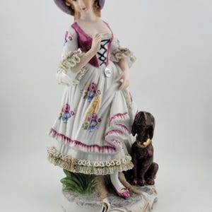 Puede incluir: Figura de porcelana de una mujer con vestido blanco con detalles florales, sombrero morado y perro marrón. El vestido tiene corpiño y ribete rosa. El perro tiene collar dorado. La figura está sobre una base decorativa.