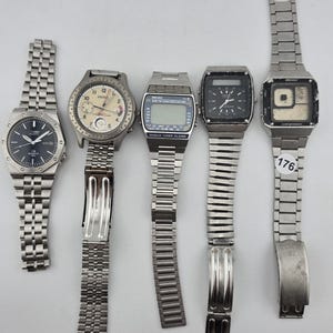 May include: Five vintage wristwatches with silver metal straps are displayed. The watches have various face designs, including analog and digital displays. One watch has a black face with a date window, while another has a digital display with the text "WORLD TIMER ALARM".