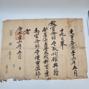 May include: A vintage, aged paper document with black calligraphy. The document features Japanese characters, a red seal, and signs of wear and tear, suggesting its age. The paper has a beige hue.