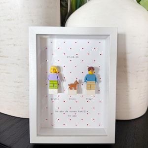 Op de afbeelding: Een witte schaduwdooslijst met twee plastic minifiguren en een klein hondje op een witte achtergrond met een rood hartjespatroon. De tekst luidt 07.25.25, Gracie, Stony, Tyler, Um ano da nossa família en Te amo.