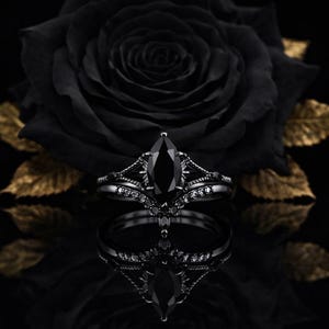 Peut inclure: Un ensemble de bagues de fiançailles noires avec une pierre noire de taille marquise. L'ensemble est posé sur un fond noir avec une rose noire et des feuilles dorées. La conception complexe de la bague comprend de petits pierres noires.