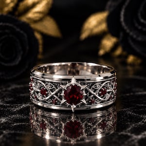 Puede incluir: Un anillo de banda en tono plateado con un intrincado patrón de enredaderas entrelazadas y pequeñas piedras rojas incrustadas. Una piedra roja redonda más grande está engastada en un marco de metal puntiagudo en forma de estrella.