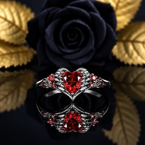 May include: A dark metal ring featuring a central heart-shaped red gemstone flanked by metallic wing-like details and smaller red accent stones along the band. The design incorporates intricate textures and a polished finish on the dark metal band.