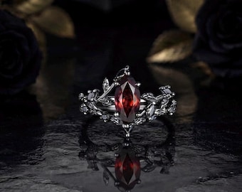 Könnte beinhalten: Ein Ring aus schwarzem Metall mit einem zentralen, tiefroten Edelstein im Marquise-Schliff. Die Ringschiene ist mit detaillierten, skulpturalen Blatt- und Rankenmotiven verziert, die vom Mittelstein ausgehen und ein botanisches Design bilden.