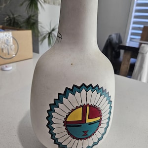 Puede incluir: Un jarrón de cerámica blanco con cuerpo redondeado y cuello estrecho. El jarrón presenta un diseño geométrico colorido con detalles turquesa, amarillos, rojos y negros. La base es negra.