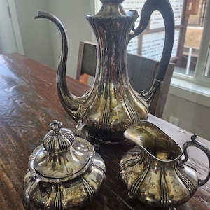 May include: An antique silver tea set, including a tall teapot with a dark handle, a sugar bowl with a lid, and a creamer. The set is displayed on a wooden table, showcasing its ornate design and aged patina.