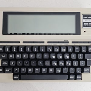May include: A vintage Radio Shack TRS-80 Model 100 portable computer. The off-white device has a black keyboard with many keys, a large LCD screen, and the Radio Shack logo. The model number is visible on the top right.