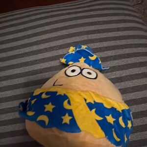 May include: A pear-shaped plush toy with large eyes and a small nose, wearing a blue hat and cape. The cape is decorated with yellow stars and crescent moons. The toy is tan with a yellow and blue cape.