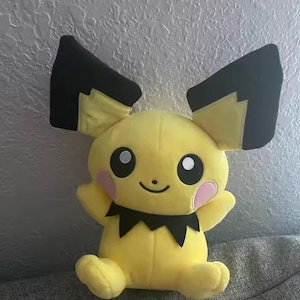 May include: A yellow plush toy with large black-tipped ears, pink cheeks, and a black zig-zag collar. The toy has a smiling face with black eyes and is sitting on a gray surface. The plush toy is a character from the popular Pokemon franchise.