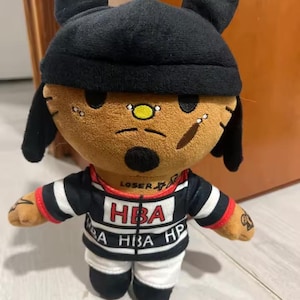 May include: A plush doll with a tan face, black hat, and black and white striped shirt. The doll has a yellow nose, facial piercings, and tattoos on its arms. The shirt has the letters "HBA" printed on it.