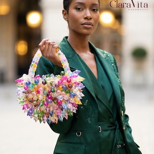 Può includere: Una borsa a mano colorata con perline, con una moltitudine di perline dai colori gioiello. La borsa è tenuta da una persona che indossa un blazer verde scuro. Il logo Clara Vita è visibile nell'angolo in alto a destra.
