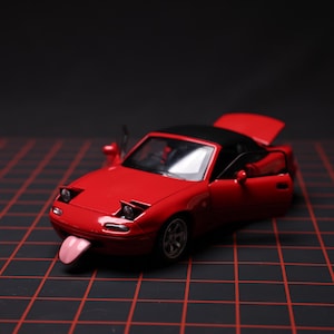 Puede incluir: Un coche de juguete rojo con techo negro, puertas abiertas y una lengua fuera. El coche tiene los faros abiertos y está sobre una superficie de rejilla negra.