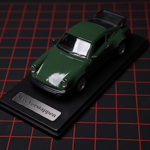Puede incluir: Un coche de modelo verde oscuro con una base negra y el texto "Max Verstappen" en una placa plateada. El coche tiene un diseño detallado, incluyendo faros y un alerón trasero. El modelo se presenta sobre una base rectangular negra.