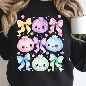 May include: A black long-sleeve sweatshirt featuring a graphic print of four colorful, smiling dumpling characters in purple, pink, blue, and mint green, interspersed with five pastel-colored bows and small floral accents and stars.