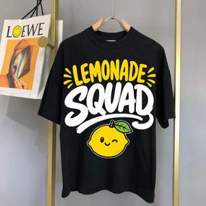 May include: Black t-shirt with the words "LEMONADE SQUAD" in yellow and white, and a smiling yellow lemon graphic. The shirt is hanging on a metal hanger against a neutral background.