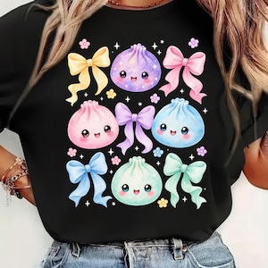 May include: A black t-shirt featuring a graphic print of four smiling, round, pastel-colored dumplings in purple, pink, blue, and mint green, interspersed with five colorful bows in yellow, pink, purple, blue, and mint green, plus small flowers.
