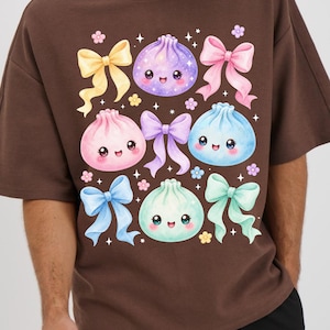 May include: A brown t-shirt featuring a graphic print of four smiling, round dumplings in purple, pink, blue, and mint green. The design includes several colorful bows in yellow, pink, purple, blue, and mint, accented by small flowers and stars.