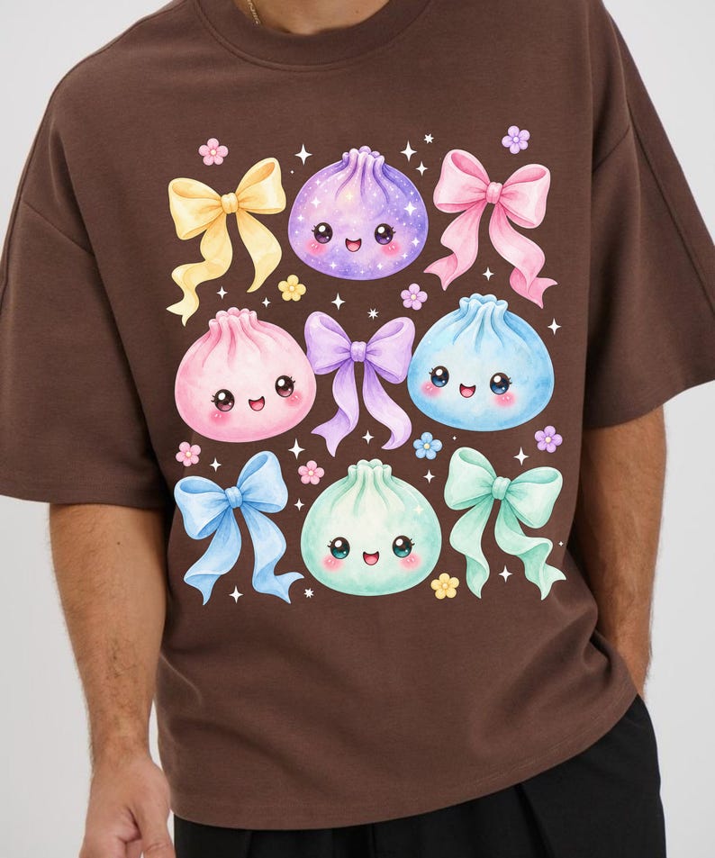 May include: A brown t-shirt featuring a graphic print of four smiling, round dumplings in purple, pink, blue, and mint green. The design includes several colorful bows in yellow, pink, purple, blue, and mint, accented by small flowers and stars.
