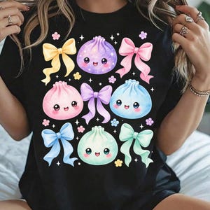 May include: A black t-shirt featuring a graphic print of four pastel-colored, smiling dumpling characters in purple, pink, blue, and mint green. The design is decorated with five colorful ribbons in yellow, pink, purple, blue, and mint, plus small flowers and stars.