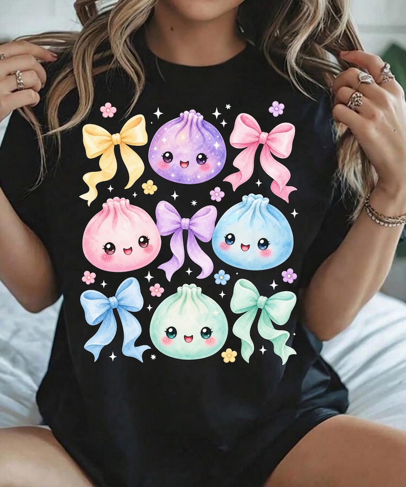 May include: A black t-shirt featuring a graphic print of four pastel-colored, smiling dumpling characters in purple, pink, blue, and mint green. The design is decorated with five colorful ribbons in yellow, pink, purple, blue, and mint, plus small flowers and stars.