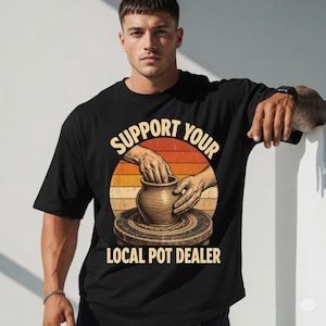May include: A black short-sleeve t-shirt featuring a graphic of hands shaping a clay pot on a pottery wheel. The design includes a sunset-colored circular background and the text "SUPPORT YOUR LOCAL POT DEALER" in a bold, yellow, sans-serif font.