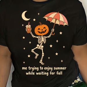 Könnte beinhalten: Ein schwarzes T-Shirt mit einer Grafik eines Skeletts mit Kürbiskopf, das einen Eiskaffee und einen gestreiften Regenschirm hält. Das Design enthält eine Mondsichel, Sterne und den Text: me trying to enjoy summer while waiting for fall.