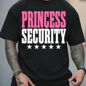 May include: A black short-sleeved t-shirt featuring the text PRINCESS in pink and SECURITY in white, followed by a row of five white stars centered on the chest. The garment has a crew neckline and a standard fit, displaying clear, bold lettering.