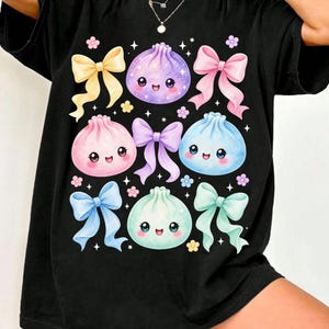 May include: A black t-shirt featuring a graphic print of four colorful, smiling dumpling characters in purple, pink, blue, and mint green. The design is decorated with various pastel-colored ribbons, small flowers, and white star accents scattered throughout.
