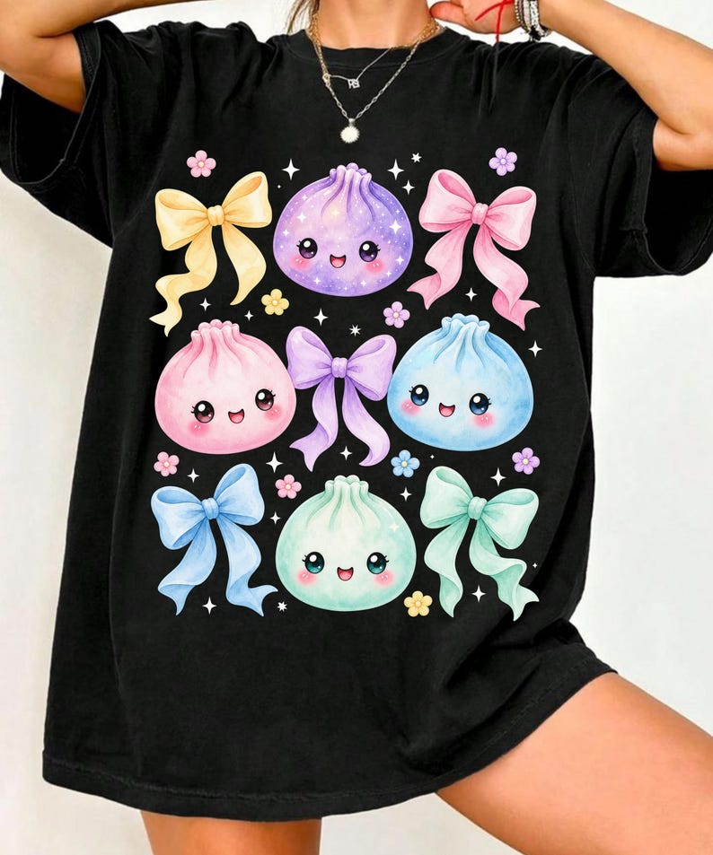 May include: A black t-shirt featuring a graphic print of four colorful, smiling dumpling characters in purple, pink, blue, and mint green. The design is decorated with various pastel-colored ribbons, small flowers, and white star accents scattered throughout.