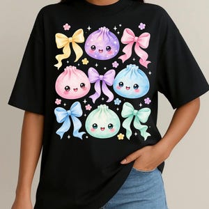 May include: A black short-sleeved t-shirt featuring a graphic print of four colorful, smiling dumpling characters in purple, pink, blue, and mint green, surrounded by pastel-colored ribbons, small flowers, and sparkling star accents.