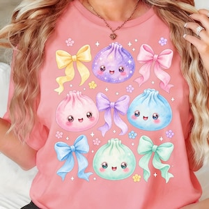May include: A coral-colored t-shirt featuring a graphic print of four smiling, round dumplings in purple, pink, blue, and mint green. The design includes five decorative bows in yellow, pink, purple, blue, and mint green, along with small flowers.
