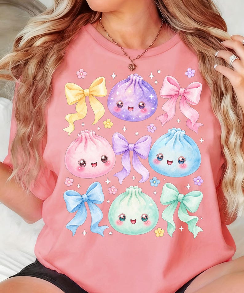 May include: A coral-colored t-shirt featuring a graphic print of four smiling, round dumplings in purple, pink, blue, and mint green. The design includes five decorative bows in yellow, pink, purple, blue, and mint green, along with small flowers.
