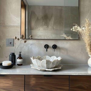 May include: A white marble vessel sink shaped like a lotus flower sits on a countertop. The bathroom features a rectangular mirror, a wall-mounted black faucet, a black soap dispenser, a black dish with a bar of soap, and a vase with dried flowers.