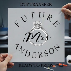 May include: A white DTF transfer sheet featuring the black text FUTURE MRS ANDERSON arranged in a circular pattern around a central illustration of a silver diamond engagement ring. The words READY TO PRESS appear at the bottom of the sheet.