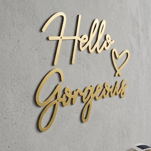 Może przedstawiać: Złota dekoracja ścienna z napisem "Hello Gorgeous" i symbolem serca. Grafika jest zamontowana na szarej ścianie w pobliżu okna. Otwarta książka leży na małym drewnianym stołku.