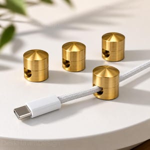 May include: Four cylindrical brass cable organisers with a brushed metallic finish. Each piece features a rounded top and a side cutout designed to hold a white braided charging cable, which is shown threaded through one of the organisers.