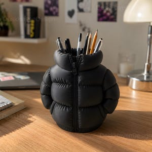 Puede incluir: Un portalápices negro con forma de chaqueta acolchada en miniatura, con cremallera central, cuello alto y paneles horizontales acolchados. El recipiente está lleno de varios bolígrafos y lápices, sobre una superficie plana.
