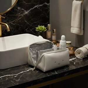 May include: A white cosmetic bag with a bunny face design, open to reveal multiple compartments. The bag is on a black marble countertop next to a white sink and various skincare products.