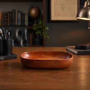 May include: An oval-shaped wooden bowl with a smooth, polished finish and a rich, warm brown grain pattern. The bowl features deep, curved sides and a flat base, designed as a functional container for small items on a desk or tabletop surface.