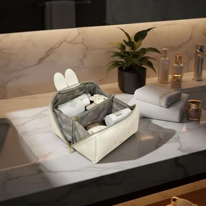 May include: A cream-coloured cosmetic bag with bunny ear accents, open to reveal toiletries. The bag is on a marble countertop, with a plant, towels, and other beauty products in the background.