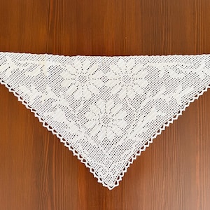 May include: A triangular white crochet shawl featuring a detailed floral pattern with three prominent daisy-like flowers. The textile is crafted with a fine mesh stitch and finished with a delicate scalloped edge along the entire perimeter.