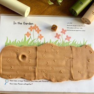 Può includere: Un set di attività di apprendimento con una base di pasta marrone, un dado e rulli numerici. Il set include un foglio di lavoro con il testo "In the Garden" e domande sui fiori. Lo sfondo è una superficie di legno.
