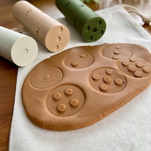 May include: A set of three cylindrical, pastel-colored wooden toys with various patterns and a brown, textured play mat. The toys are light green, tan, and olive green. The play mat has circular indentations with textured details.