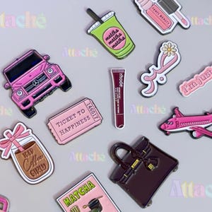 May include: A collection of enamel pins featuring a pink SUV, a green matcha drink, a Fenty Beauty lip product, a pink flower, a pink ticket labelled Ticket to Happiness, a rhode lip tube, a pink aeroplane, an iced coffee drink, and a dark handbag.