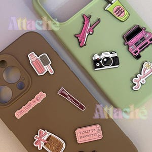 May include: Two smartphone cases in sage green and brown, decorated with various enamel pins. The green case features a pink aeroplane, a pink SUV, a black camera, a green matcha drink, and a pink flower. The brown case displays a pink lip gloss, a tube labelled rhode, a ticket labelled TICKET TO HAPPINESS, a coffee cup, and text reading i'm just a girl.