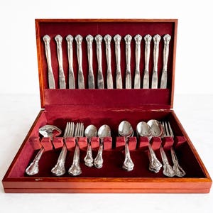 May include: A complete set of silver silverware, including knives, forks, and spoons, is neatly arranged in a wooden case with a red velvet lining. The silverware features ornate handles and is designed for formal dining occasions.
